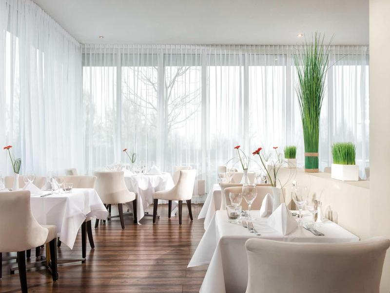 Helles Restaurant mit weißen Stühlen, Tischdecken, Holzfußboden und großen Fenstern mit Vorhängen.