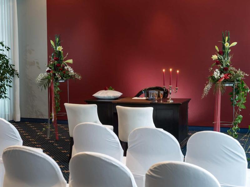 Weiß gedeckte Stühle vor roter Wand, Tisch mit Kerzen und Blumenarrangements rechts und links.