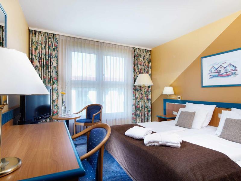 Helles Hotelzimmer mit Doppelbett, Schreibtisch, Stühlen, Teppichboden und großen Fenstern mit Vorhängen.