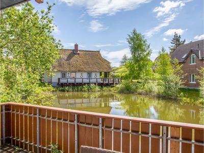Blick von einem Balkon auf einen Teich mit Häusern und grünem Laub unter blauem Himmel.