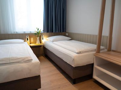 Twin room with simple beds, bedside table, plants, and curtain at the window.