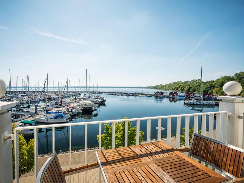 Blick von Balkon mit Holztisch auf Hafen mit Segelbooten und blauem Himmel.