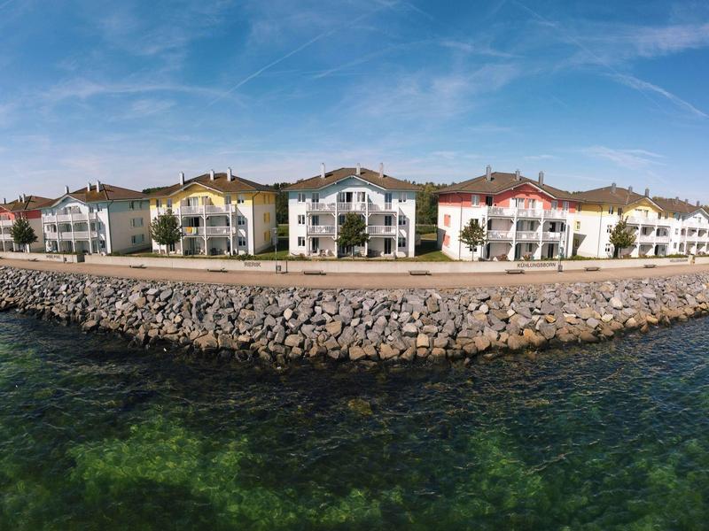 Reihe weißer Häuser mit roten Dächern am Ufer, davor klare grüne Meereswellen und blauer Himmel.