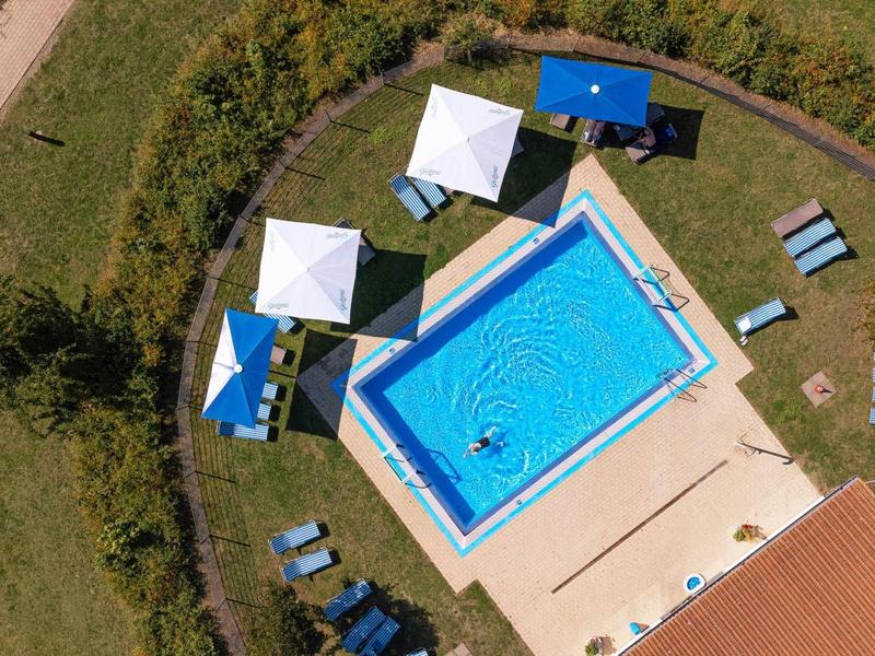 Ein rechteckiges Schwimmbecken mit blauen Wasserspritzern, umgeben von Sonnenschirmen und Liegestühlen.