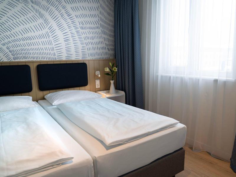 Helles Schlafzimmer mit zwei Einzelbetten, weißer Bettwäsche, blauen Kopfkissen und Vorhängen.