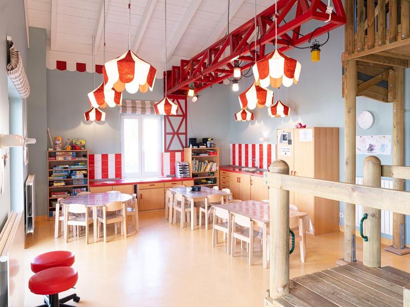 Heller, freundlicher Kindergartenraum mit roten Akzenten, Holzmöbeln und hängenden Laternenlampen.