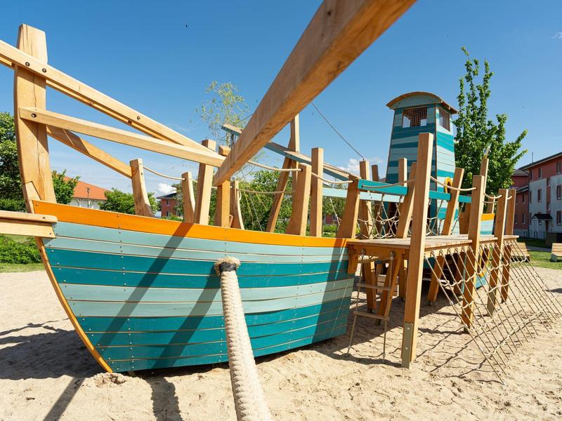 Holzspielplatz in Form eines Schiffes mit blau-grünen Streifen auf Sandboden unter blauem Himmel.