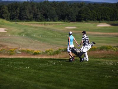 Zwei Personen gehen mit Golfbags über einen grünen Golfplatz mit Bäumen im Hintergrund.