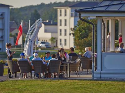 Menschen sitzen im Außenbereich eines Cafés mit Blick auf Grünfläche und Gebäude im Hintergrund.