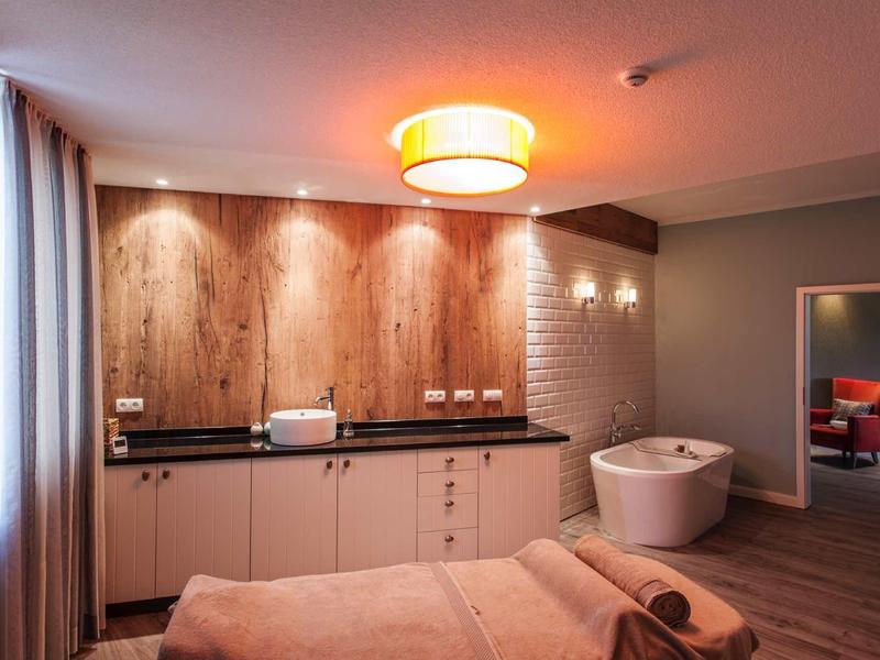 Gemütliches Schlafzimmer mit Holzwand, Badewanne, warmem Licht und hellen Vorhängen.