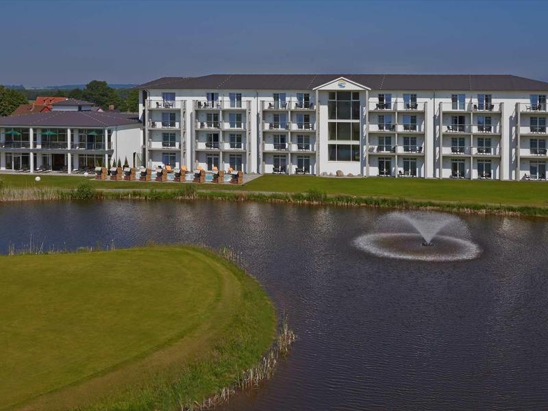 Mehrstöckiges Wohngebäude mit weißen Balkonen am See mit Springbrunnen unter bewölktem Himmel.