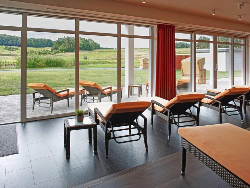 Moderner Raum mit großen Fenstern, Liegestühlen und Blick auf grüne Landschaft.