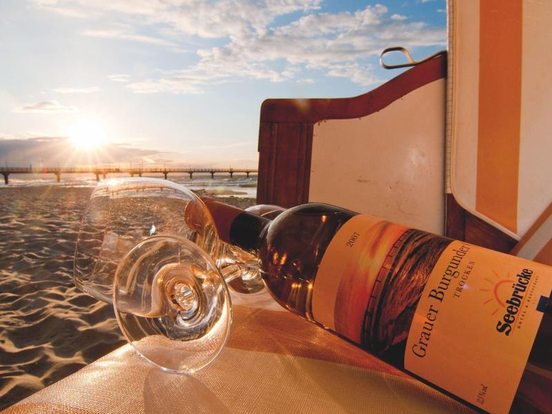 Weinflasche und Glas liegen am Strand mit Blick auf den Sonnenuntergang und eine Promenade.