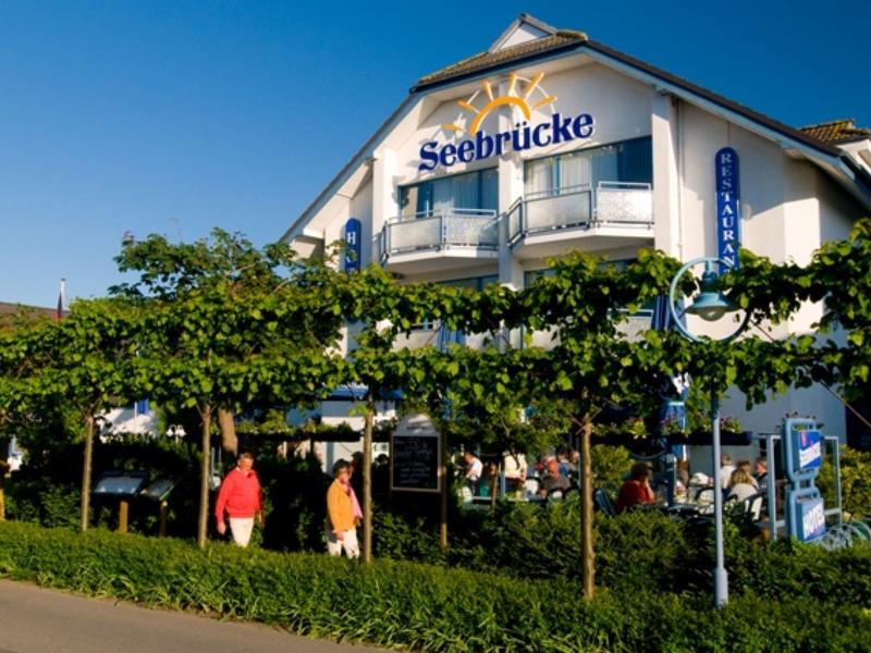 Ein weißes Gebäude mit dem Schriftzug "Seebrücke" unter blauem Himmel, davor grüne Pflanzen.