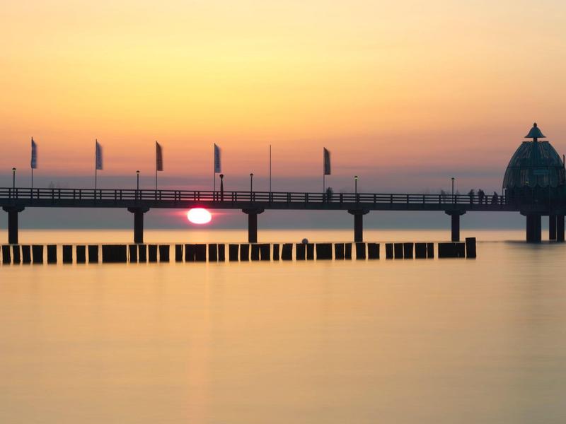 Sonnenuntergang über einer langen Seebrücke mit Menschen und einem gläsernen Pavillon am Ende.
