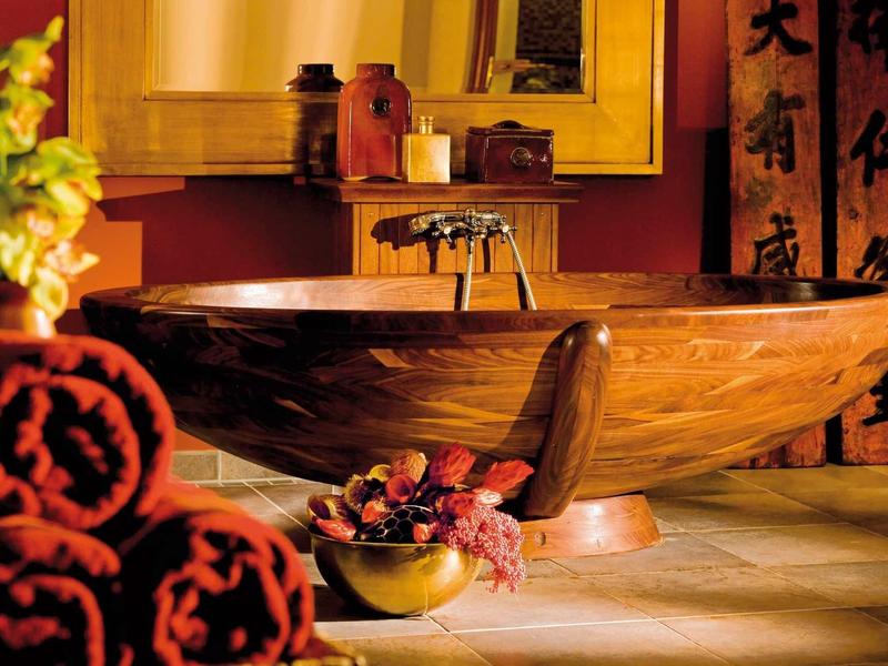 Rustikale Holzbadewanne in warmem Licht, rote Handtücher, Pflanzen und asiatische Deko im Hintergrund.