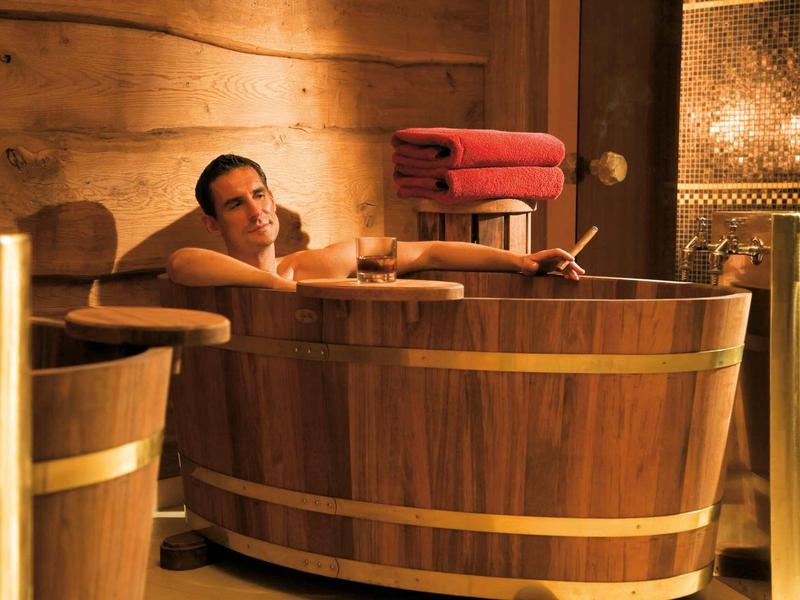 Mann entspannt in einem Holzzuber in einem warm beleuchteten, rustikalen Spa-Raum.