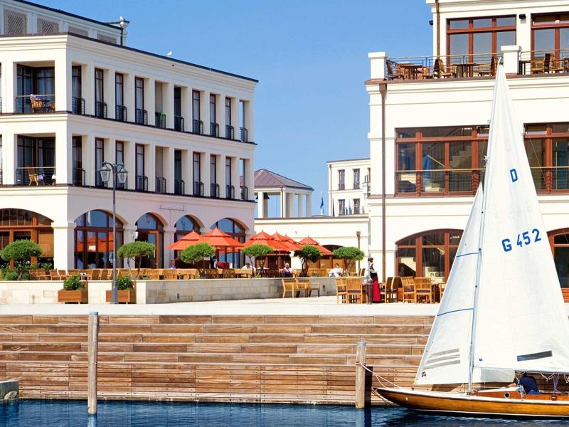 Segelboot vor weißem Gebäude mit Terrassen und orangefarbenen Sonnenschirmen am Wasser.
