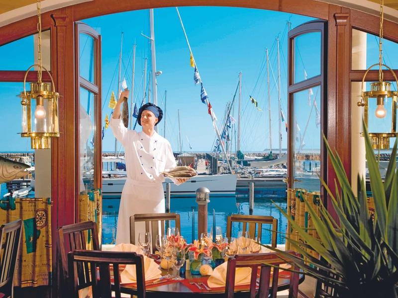 Blick durch offene Fenstertüren auf Terrasse mit gedecktem Tisch, Kellner und Hafen im Hintergrund.