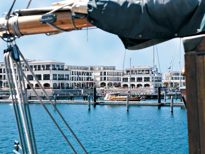 Hafen mit blauem Wasser, Segelschiffmast im Vordergrund und moderner Gebäudereihe im Hintergrund.