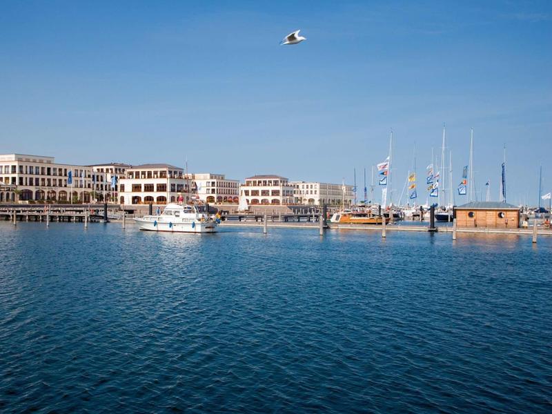 Hafen mit Segelbooten und Gebäuden, blauer Himmel, ruhiges Wasser und Möwe in Flug.