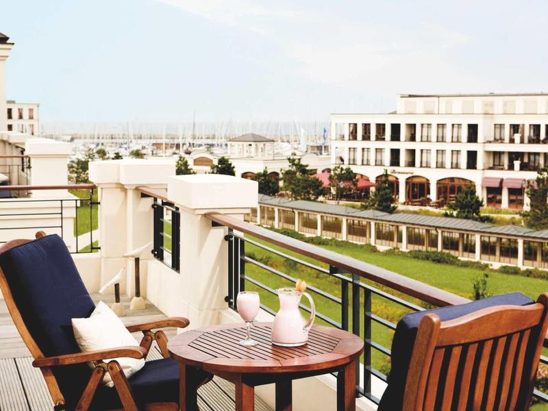 Balkon mit Holzstuhl, Tisch, Teekanne, Pflanzen und Blick auf moderne Gebäude und Wasser im Hintergrund.