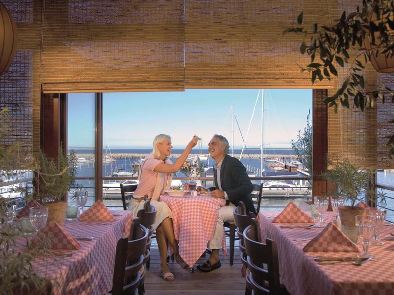 Zwei Personen sitzen im Restaurant mit rot-weiß karierten Tischdecken, Meerblick und warmem Licht.