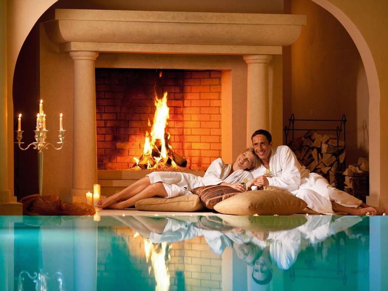 Paar entspannt auf Kissen vor brennendem Kamin neben blauem Hallenbad mit Kerzenlicht.