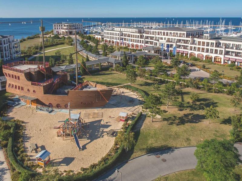 Grüner Park mit piratenschiffförmigem Spielplatz, umgeben von modernen weißen Gebäuden am Meer.
