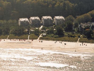 Strand met enkele gebouwen aan de rand van een bos en mensen langs de kust op een zonnige dag.