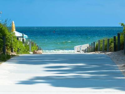 Blick auf einen breiten Weg zum Meer, umgeben von Sand, Grünpflanzen und blauem Himmel.