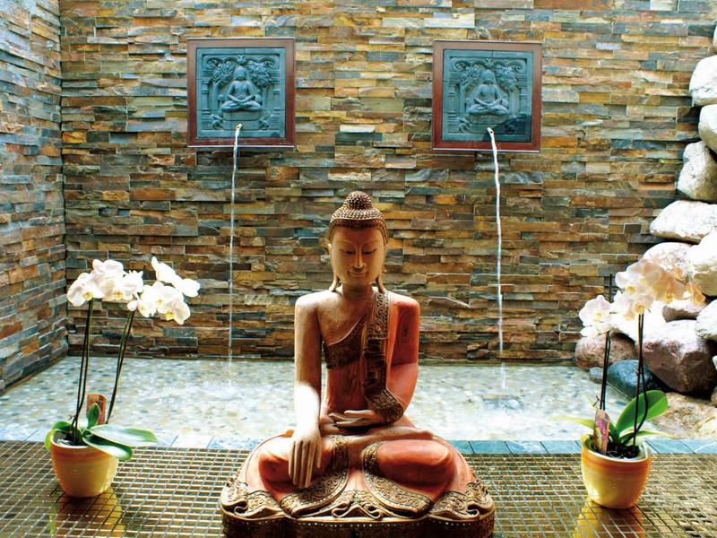 Een Boeddhabeeld zit tussen twee orchideeën voor een stenen muur met waterfonteinen.
