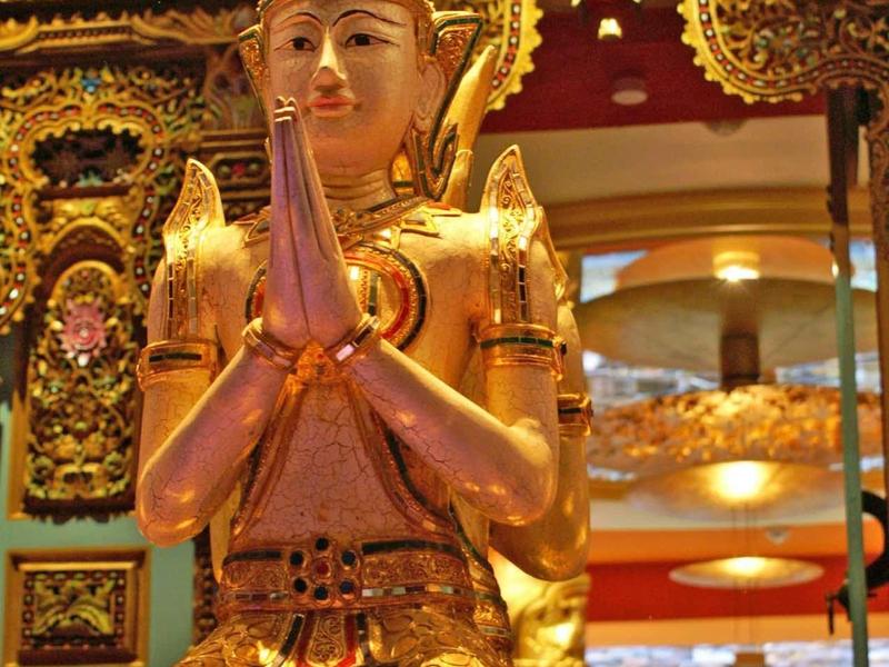 Gouden beeld met gevouwen handen voor een sierlijke tempelachtergrond.