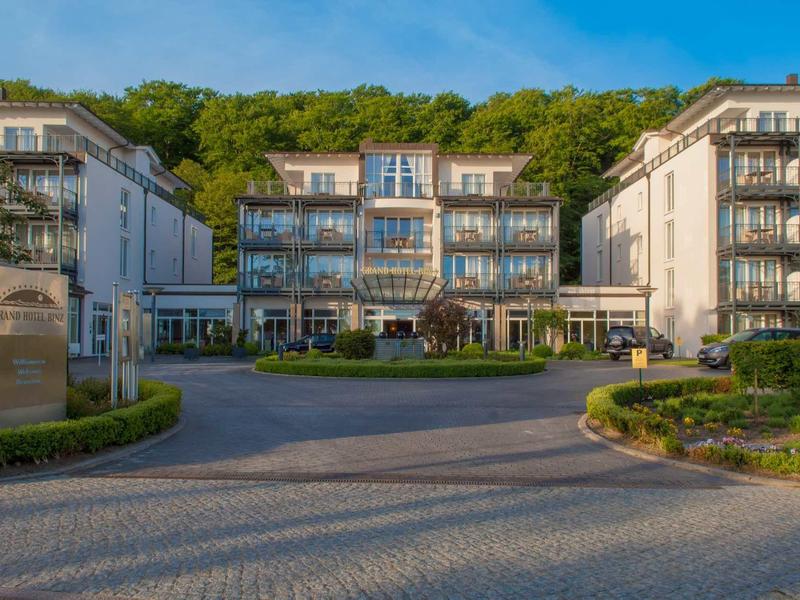 Modern hotel met drie gebouwen, groene achtergrond en geplaveide voorplaats.