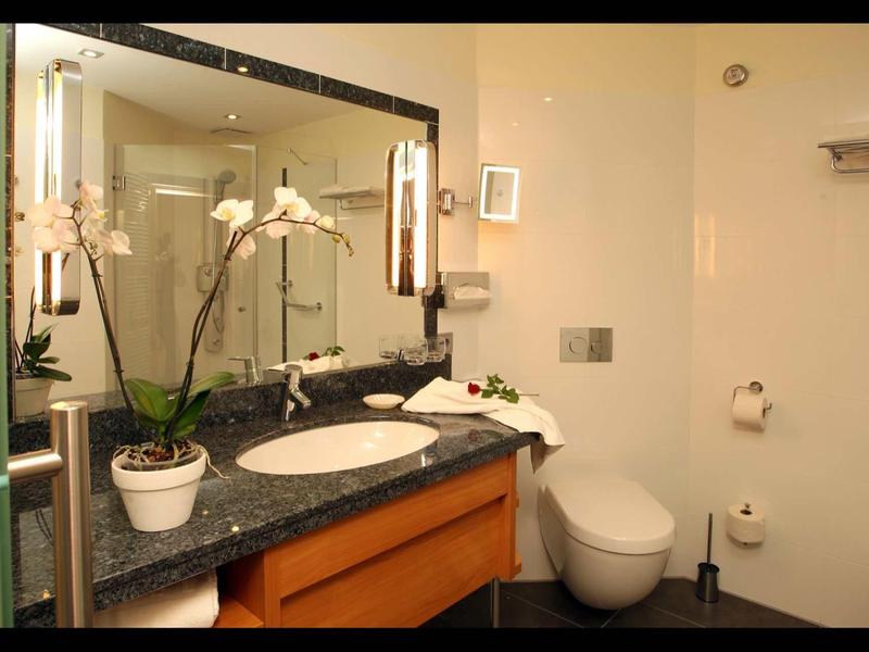 Modernes, helles Badezimmer mit Waschbecken, großer Spiegel, Blumen und WC, klare Linien, helle Wände.