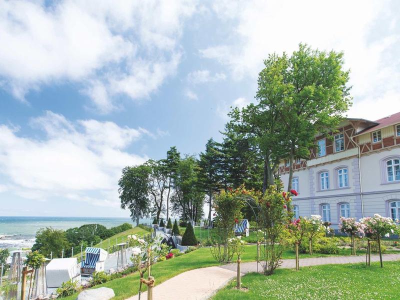 Großes Haus mit Garten und Bäumen an der Küste unter blauem Himmel mit Wolken.