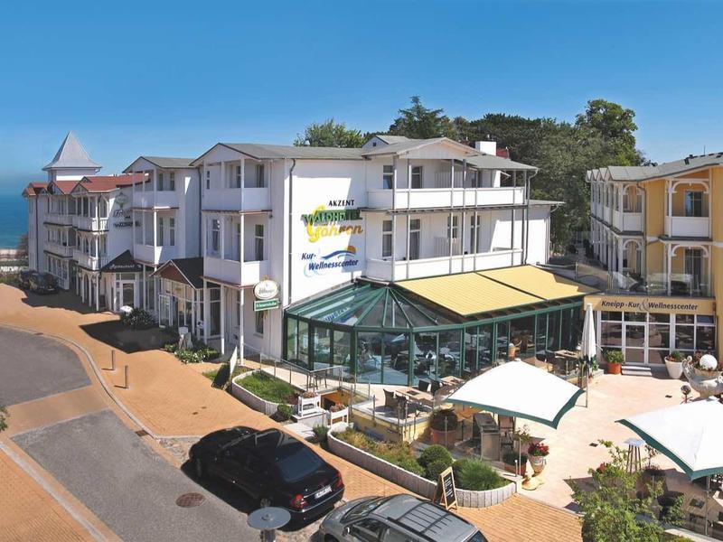 Straße mit mehrstöckigen Häusern, Autos und Cafés unter Sonnenschirmen bei Meerblick im Hintergrund.