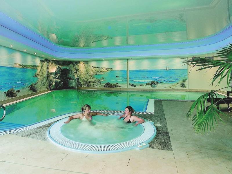 Innen-Schwimmbad mit großem Pool, runden Whirlpool, Palmen und tropischer Wandbemalung.