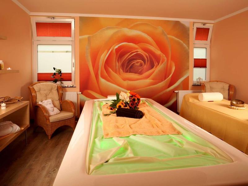 Ein warm beleuchteter Behandlungsraum mit zwei Massageliegen, einer großen Rosenwanddekoration und sanften Farben.