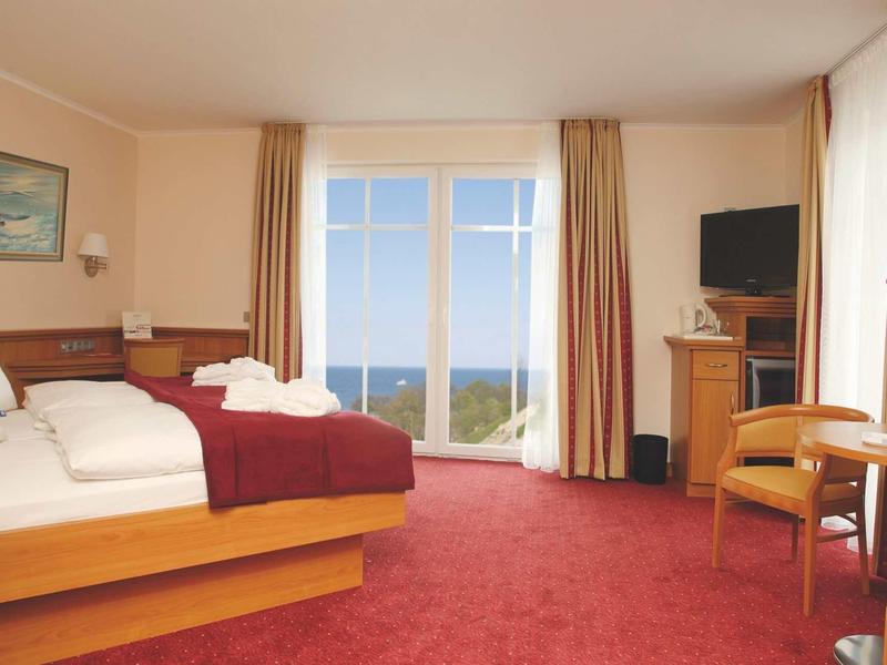 Hell eingerichtetes Hotelzimmer mit Doppelbett, rotem Teppich, TV, Tisch, Stühlen und Meerblick.