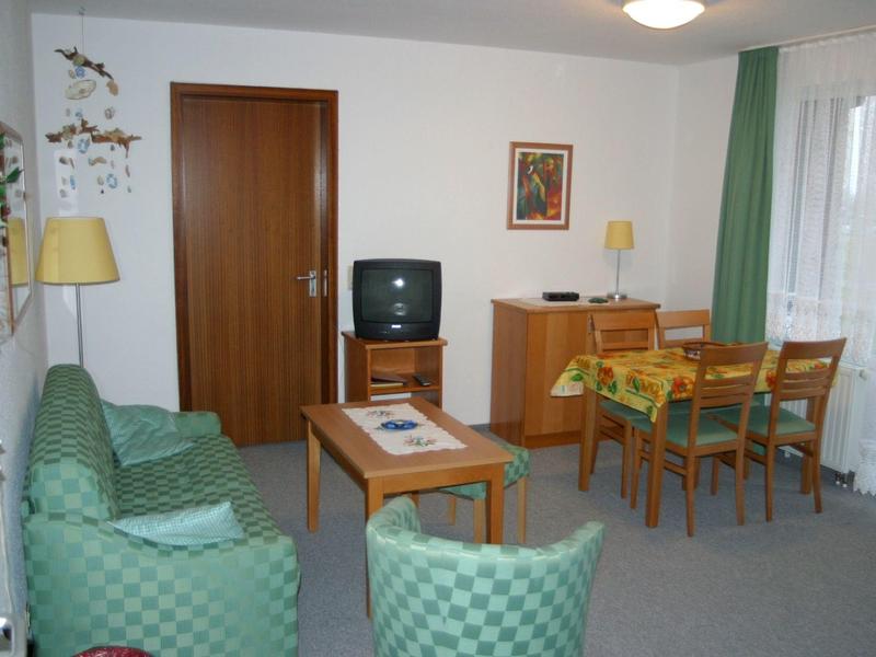 Gemütliches Wohnzimmer mit grünen Möbeln, Holztisch, Stühlen und TV vor weißer Wand.