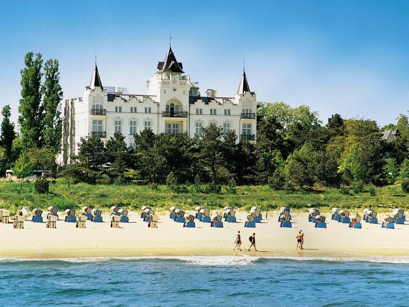 Strand mit vielen Strandkörben, davor Meer, im Hintergrund großes weißes Schloss und viele grüne Bäume.