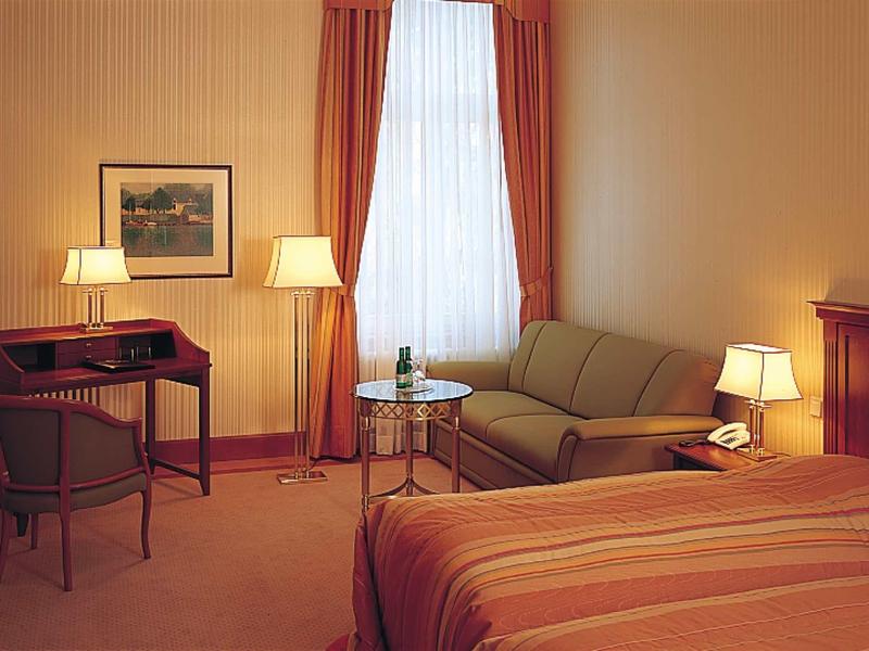 Gemütliches Hotelzimmer mit Bett, Sofa, Schreibtisch, Lampen und großem Fenster mit Vorhängen.