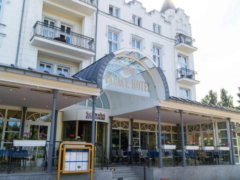 Eingang zu einem weißen Hotel mit großem Glasportal und überdachter Terrasse mit Tischen und Stühlen.