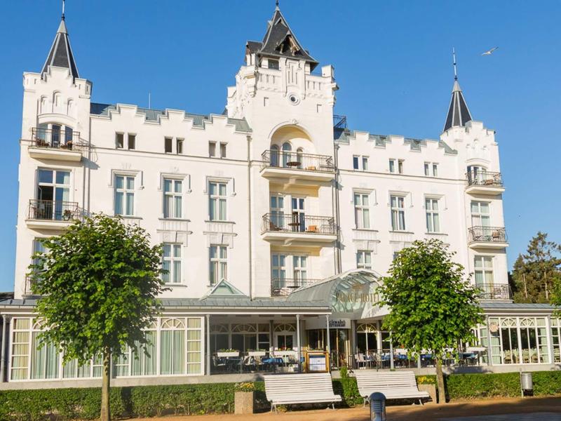 Weißes Schloss mit Zinnen und Türmen, großem Wintergarten und grünen Bäumen im Vordergrund.