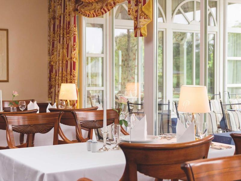 Helles Restaurant mit Holzmöbeln, weißen Tischdecken und großen Fenstern zur Terrasse.