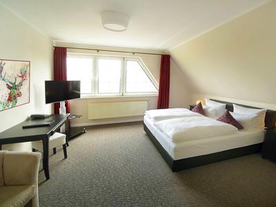 Hell eingerichtetes Hotelzimmer mit großem Bett, Sessel, Schreibtisch und Fenster mit rotem Vorhang.