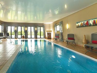 Helles Hallenbad mit großem Fensterfront, blauen Wasser und roten Liegestühlen an der Wand.