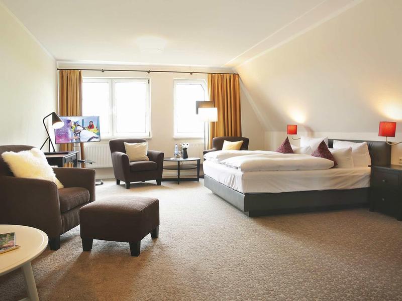 Geräumiges Hotelzimmer mit Doppelbett, zwei Sesseln, Teppichboden und großen Fenstern.