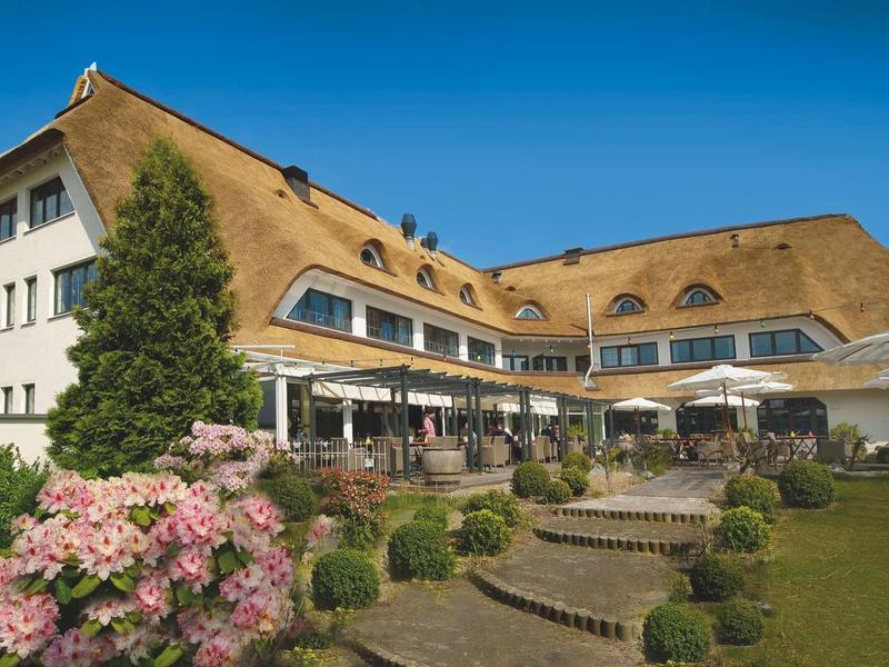Großes Fachwerkhaus mit strohgedecktem Dach, Terrasse, Blumen und blauem Himmel.